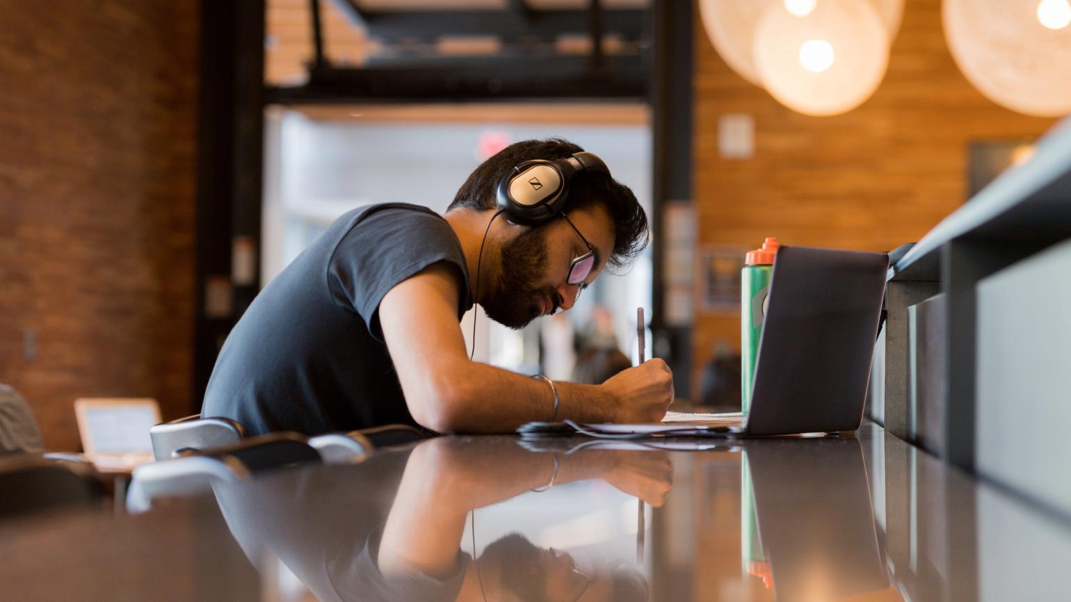 A student wearing headphones writes in a notebook with a laptop beside them.