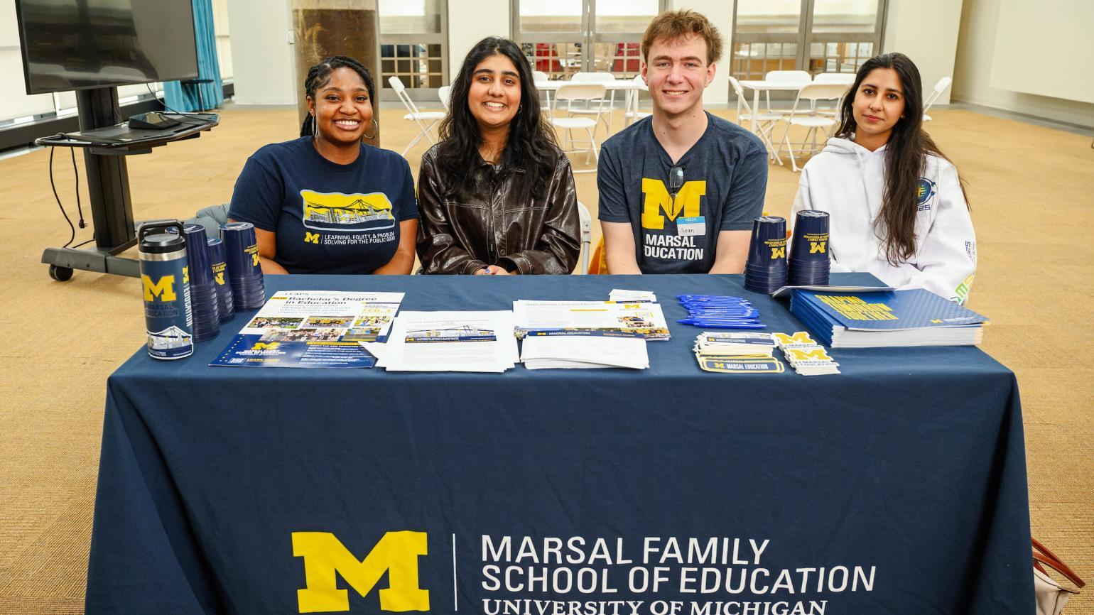 Four young people sit at a table, ready to hand out flyers and water bottles bearing the Marsal Family School of Education logo.