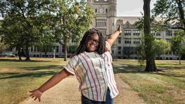 Teenager, wearing a white shirt with vertical stripes in primary colors and jeans, with long locs, shown from the waste up, with their arms outstretched, smiling at the camera. The Marygrove building's entrance towers in the background. 