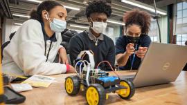 Three people wearing masks working on a laptop, with a 4-wheel robotic prototype beside them.