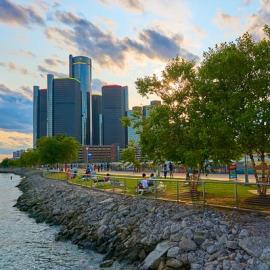 Detroit Riverwalk on a sunny day. The circular tower of the Renaissance Center dominates the background, with the sun setting behind it. In the foreground, the Detroit river meets an embankment of stones, along which groups of people walk, bike, and sit in lawn chairs.