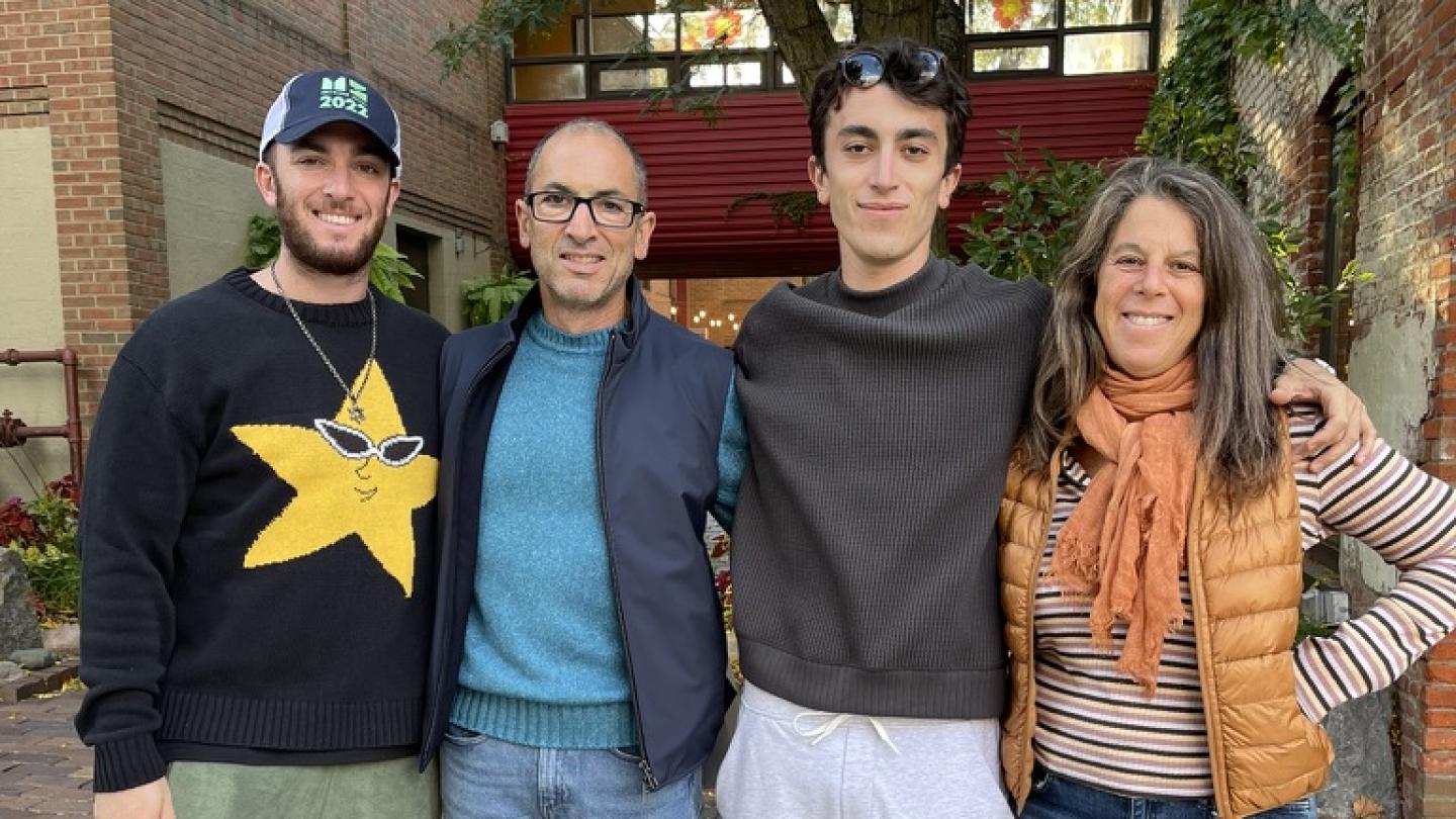A family of four—two young adult men and their parents—pose outside with their arms around each other. They stand in on a patio outside a brick building, in front of a tree.