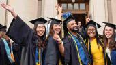 Five new graduates in commencement attire smile and pose for a photo in front of Hill Auditorium