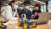 Three people wearing masks working on a laptop, with a 4-wheel robotic prototype beside them.