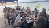 A large group of ELD interns sit at a table eating lunch in front of Lake Tequesquitengo. A blue umbrella offers shade on the visibly sunny day.