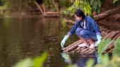 A student on a raft made of logs, wearing gloves, reaches into water.