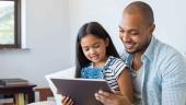 Father and smiling daughter sitting on sofa using digital tablet.