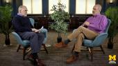 Don Peurach and Barry Fishman sit across from each other in blue armchairs, having a conversation in a formal setting with plants and large windows in the background.