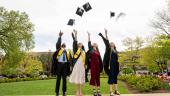 Four individuals dressed in graduation attire throw their graduation caps toward up toward a sprawling, cloudy sky.