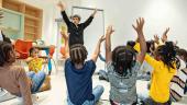 Approximately 7 young children imitate the teacher by raising both arms into the air and spreading their hands open.