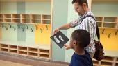 A teacher and a young student walk through a colorful hallway. The teacher is holding a piece of art made by the student.