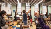 A teacher holds her thumb up in the air and approximately 6 students respond by holding their thumbs up. The room is a high school engineering classroom with electrical cords reaching from the ceiling to the work benches.