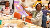 A young child proudly holds a piece of art he made depicting a bird sitting on a branch. Other children work on their art projects in the background.