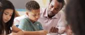 Teacher of color crouches beside student of color as they work together on a writing task